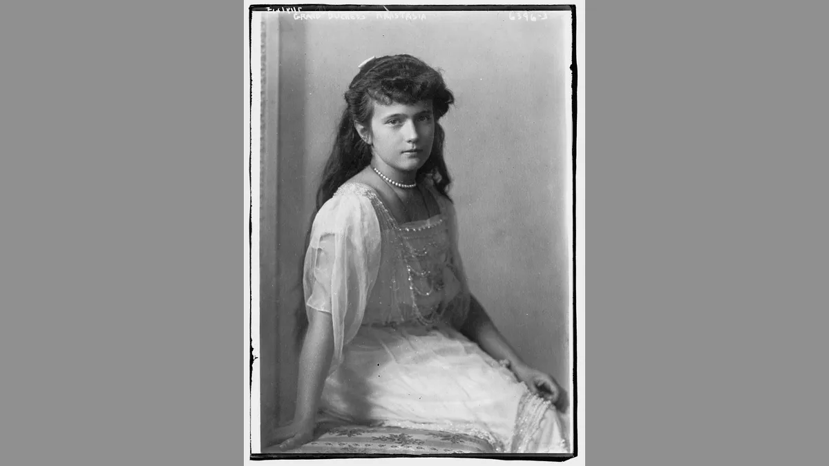 Retrato em preto e branco de uma menina de cabelo longo e escuro preso com laço, colar de pérolas e vestido claro rendado, sentada de lado e olhando para a câmera.