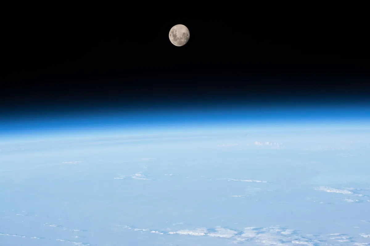Vista da Terra a partir da órbita: a superfície esbranquiçada embaixo, uma faixa azul fina no horizonte e, acima dela, o espaço negro com a Lua cheia.