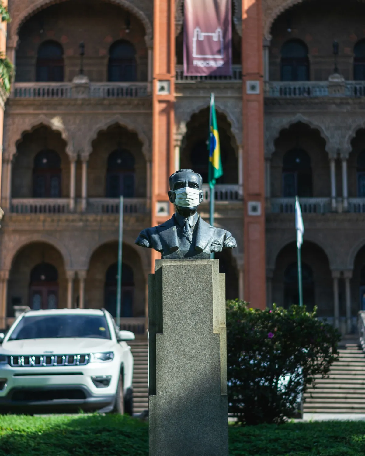 Busto de bronze de um homem de terno, com uma máscara cirúrgica branca no rosto, sobre um pedestal de granito, diante de um prédio de tijolos com arcos em estilo mourisco e uma faixa com o nome Fiocruz.