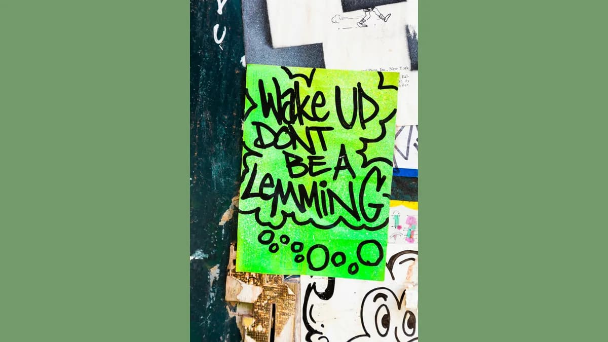 Cartaz verde-limão escrito à mão com marcador preto, colado numa parede cheia de papéis rasgados e adesivos, com balão de pensamento desenhado em volta das palavras em inglês.