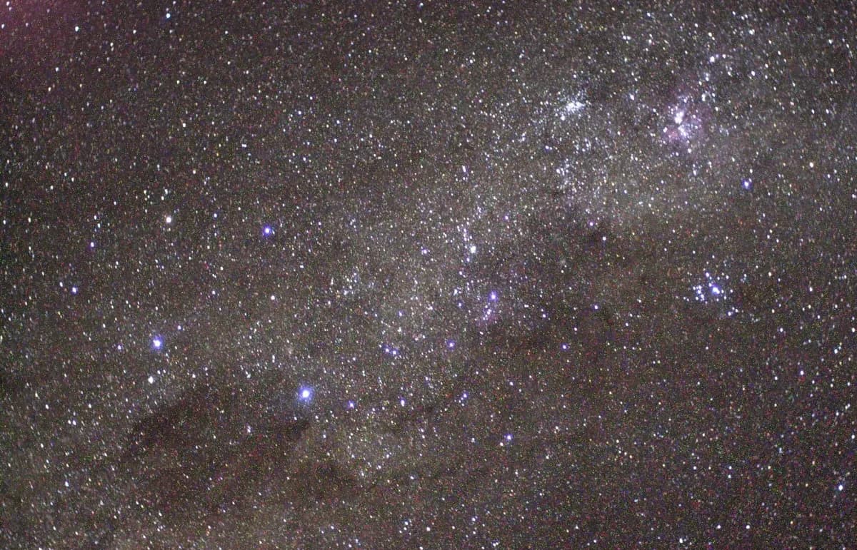 Campo denso de estrelas da Via Láctea; à esquerda, algumas estrelas azuladas mais brilhantes se destacam acima de uma faixa escura, e há uma nuvem rosada no alto à direita.