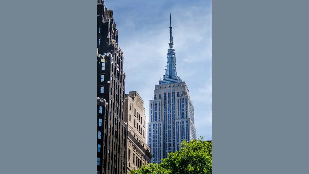 Torre do Empire State Building vista de baixo, com a antena fina no topo contra um céu azul com nuvens, entre prédios mais baixos e copas de árvores.