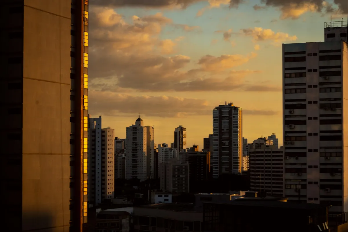 Prédios residenciais altos de Goiânia vistos do alto ao entardecer, recortados contra um céu alaranjado com nuvens.