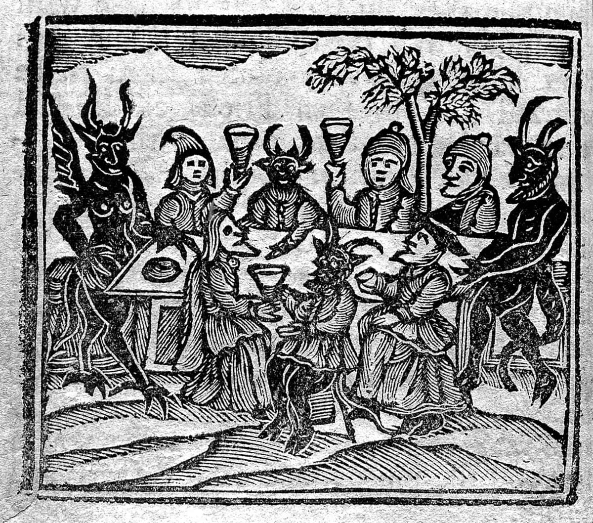 Xilogravura em preto e branco: figuras humanas de chapéu pontudo brindam com taças numa mesa ao ar livre, cercadas por demônios com chifres, asas e caudas.