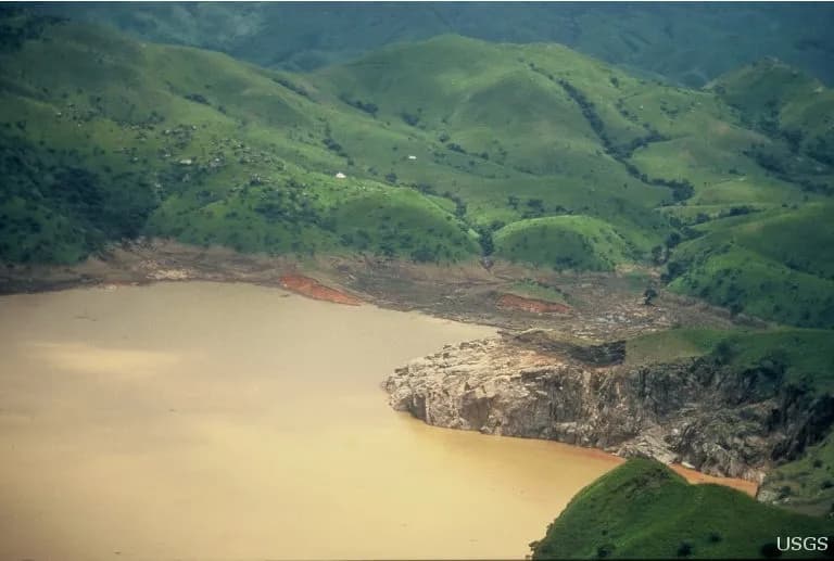 Vista do alto de um lago de água marrom-alaranjada cercado por morros verdes, com um paredão de rocha clara avançando sobre a água e as margens nuas, sem vegetação, logo acima da linha d'água.