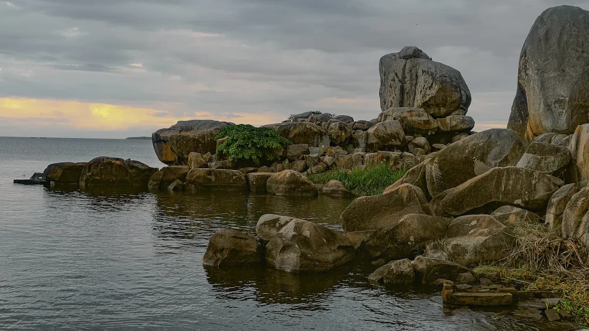Margem de lago com grandes rochas arredondadas empilhadas, um pouco de vegetação entre elas e água calma sob céu nublado, com uma faixa amarela de sol no horizonte.