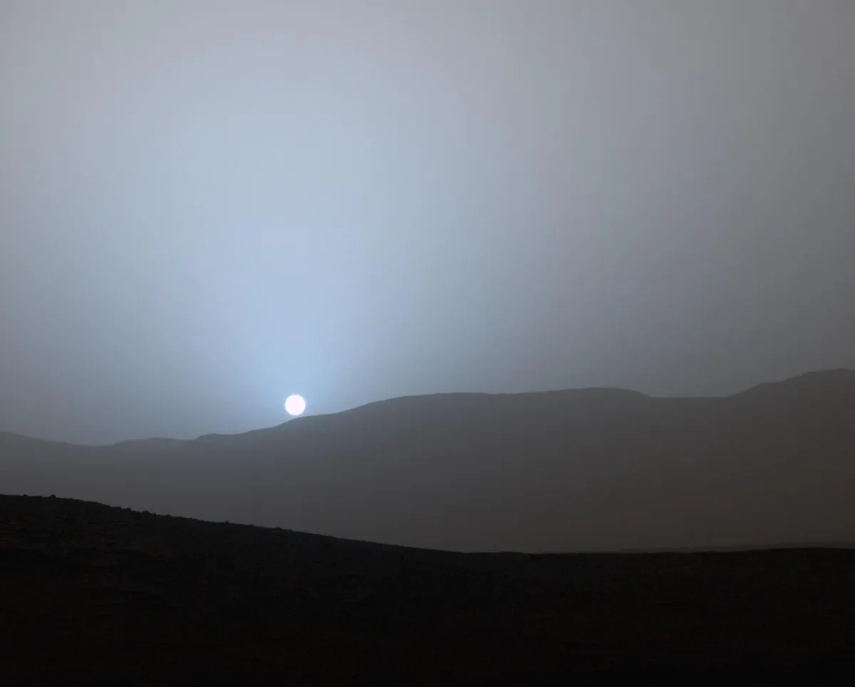 Paisagem escura e acinzentada com colinas no horizonte e um pequeno disco branco do Sol, cercado por um halo azulado no céu cinza.