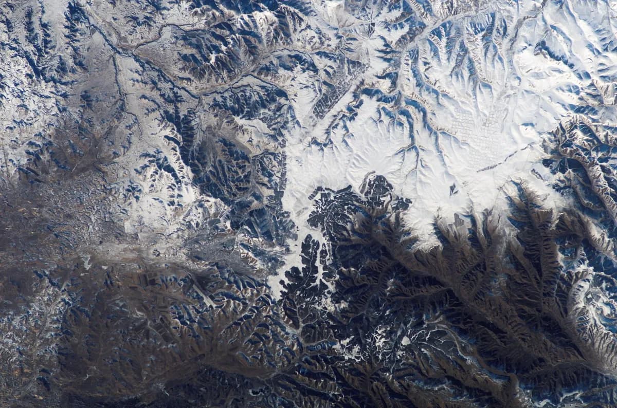 Vista do alto de montanhas e vales cobertos de neve, com encostas sombreadas em azul e marrom e um padrão quadriculado de lavouras no canto superior direito.