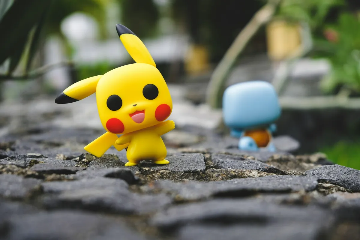 Boneco de vinil do Pikachu amarelo com bochechas vermelhas e orelhas de ponta preta sobre um calçamento de pedra, com outro boneco azul desfocado ao fundo.