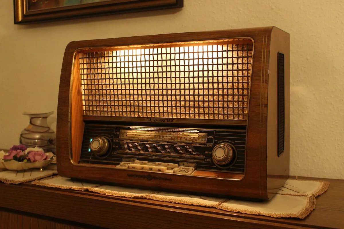Rádio de mesa com caixa de madeira escura, grade dourada iluminada por dentro e dois botões giratórios, sobre um móvel com toalhinha de crochê.