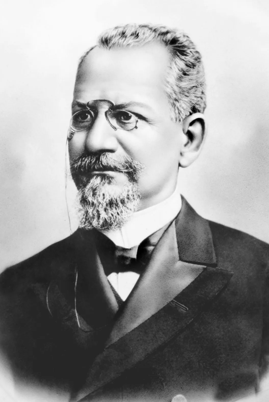 Retrato em preto e branco de um homem de cabelos grisalhos, cavanhaque, pincenê e terno escuro com gravata-borboleta.