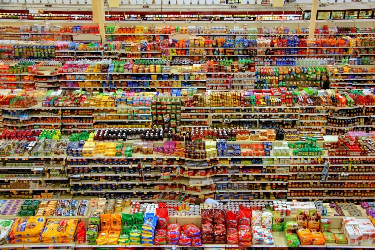Vista do alto de um supermercado com dezenas de gôndolas abarrotadas de embalagens coloridas de alimentos, de petiscos a enlatados, e um cliente entre elas ao fundo.