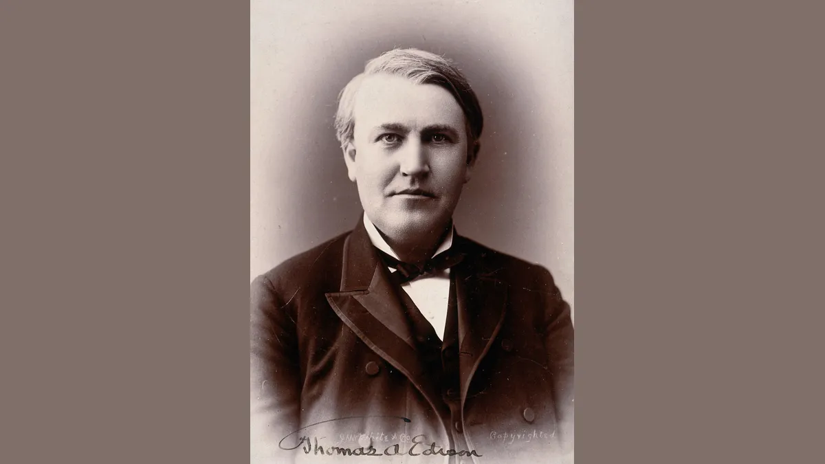 Retrato fotográfico em tom sépia de um homem de cabelos grisalhos penteados de lado, de terno escuro, colete e gravata-borboleta, com uma assinatura manuscrita na parte de baixo.