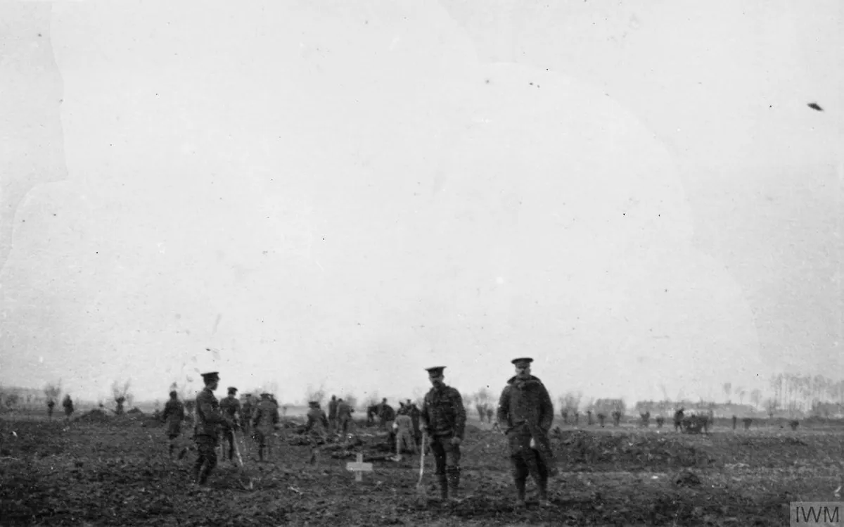 Soldados de uniforme e quepe de pé num campo de terra revirada sob céu branco; à frente, uma pequena cruz cravada no chão e um homem apoiado numa pá.