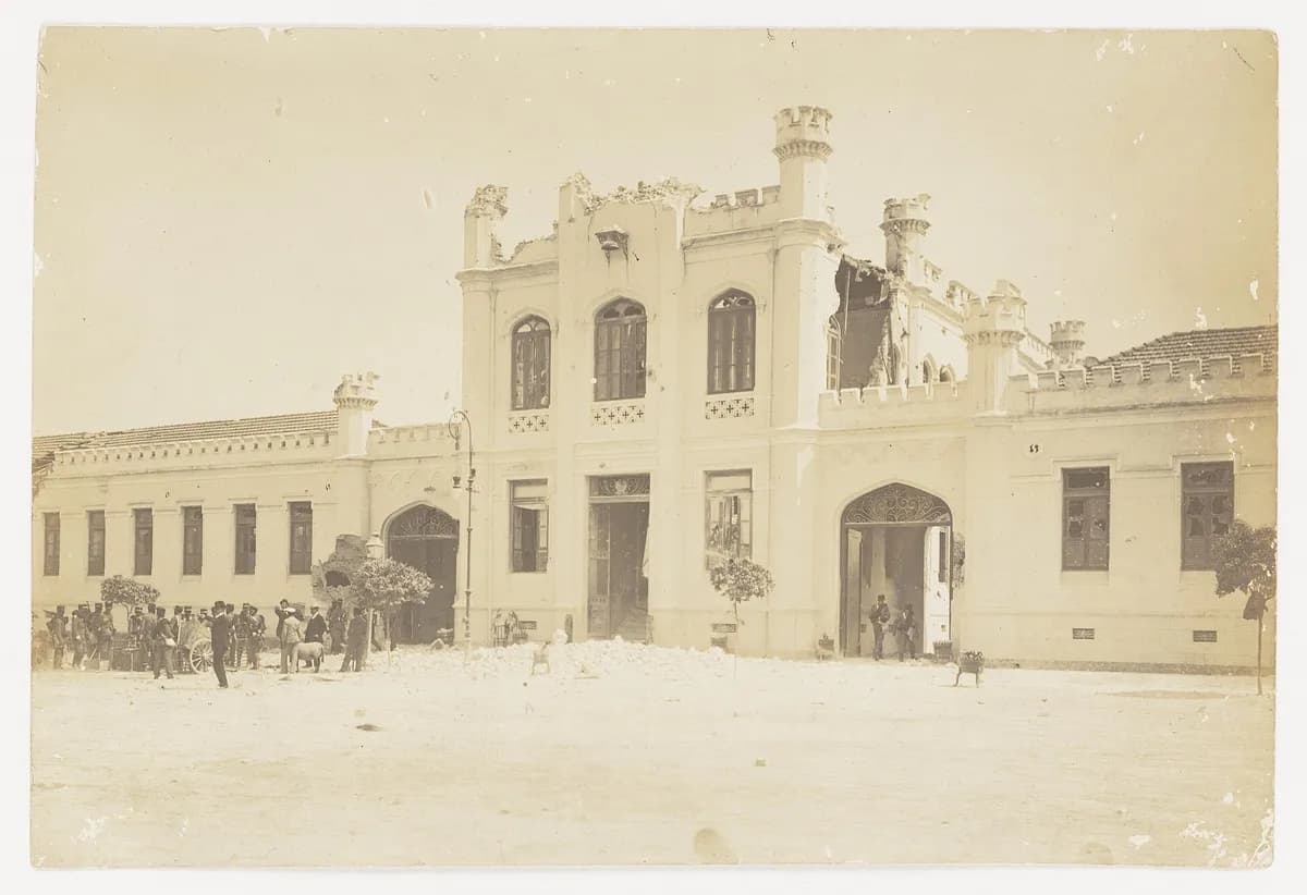 Fachada em sépia de um quartel de dois andares com ameias, com parte do topo desmoronada e entulho na frente; à esquerda, um grupo de homens de terno e uniforme.