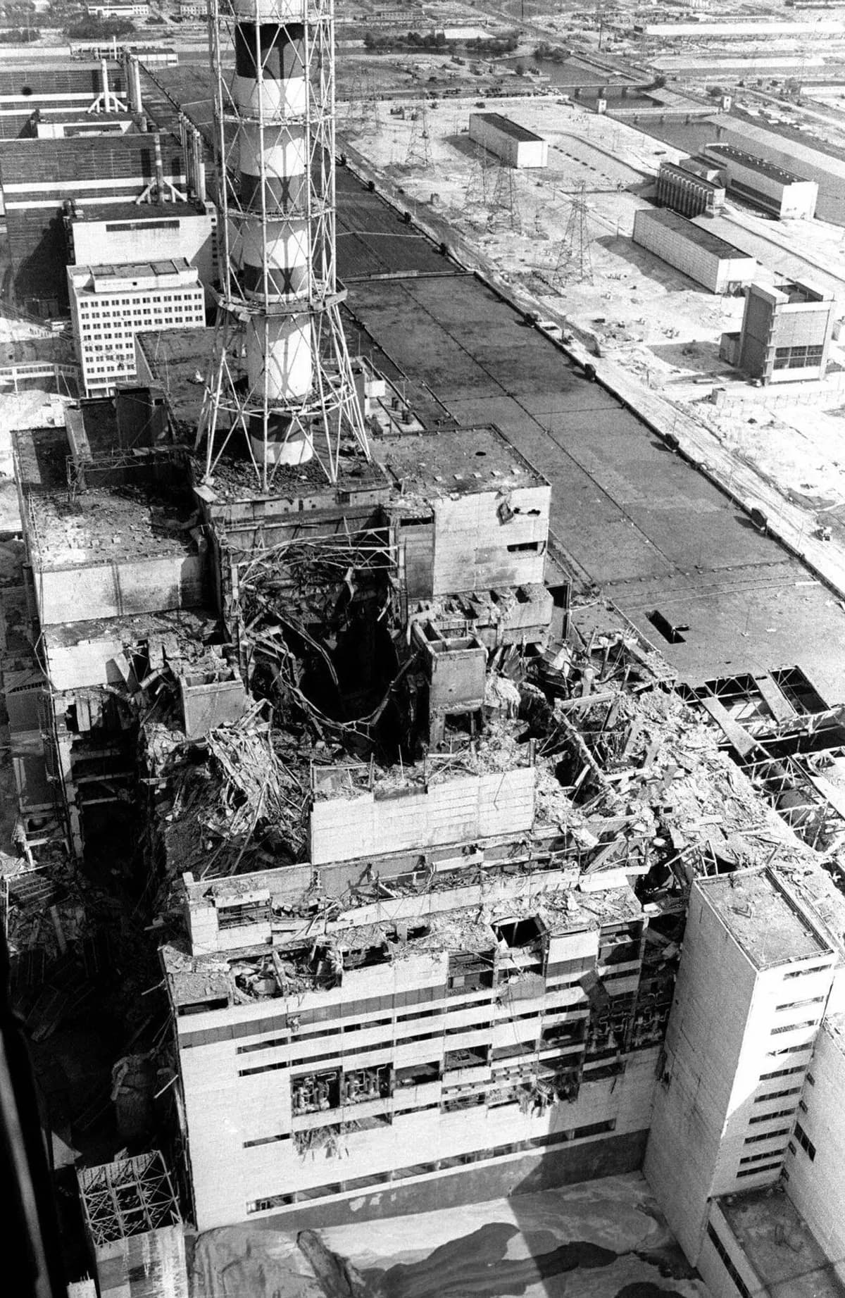 Foto aérea em preto e branco de um prédio industrial com a cobertura e as paredes arrebentadas, estruturas retorcidas expostas e uma alta chaminé listrada ao fundo.