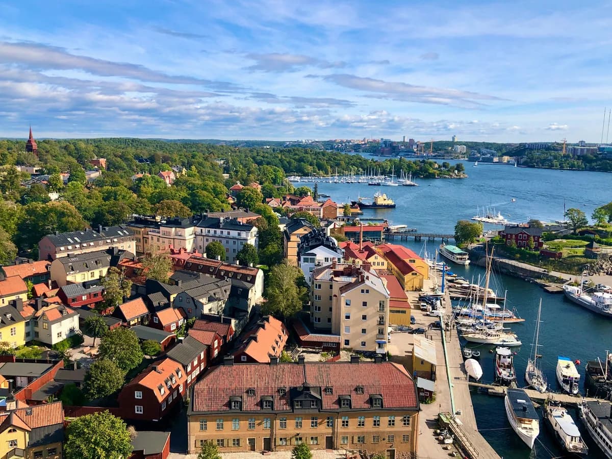Vista do alto de Estocolmo no verão, com telhados vermelhos e amarelos, árvores, um canal com barcos atracados e céu azul com nuvens.