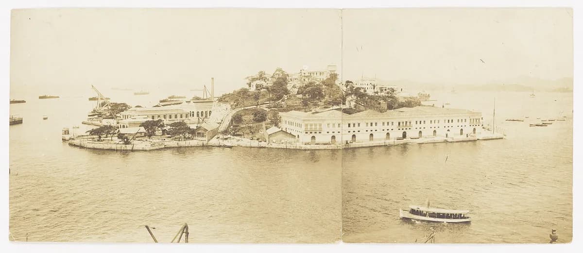 Panorama em sépia de uma ilha baixa na baía, coberta por prédios longos de dois andares e um morro arborizado no centro; um barco a vapor passa em primeiro plano.